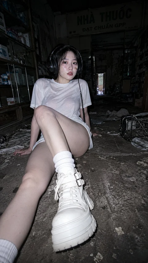 A seated figure in a light T-shirt extends a white high-top sneaker toward the camera, framed by a dark, worn room.